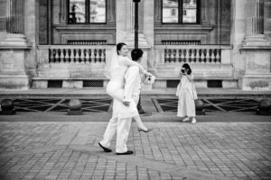 Un marié porte sa mariée sur son dos pendant qu'une photographe immortalise la scène devant la pyramide du Louvre